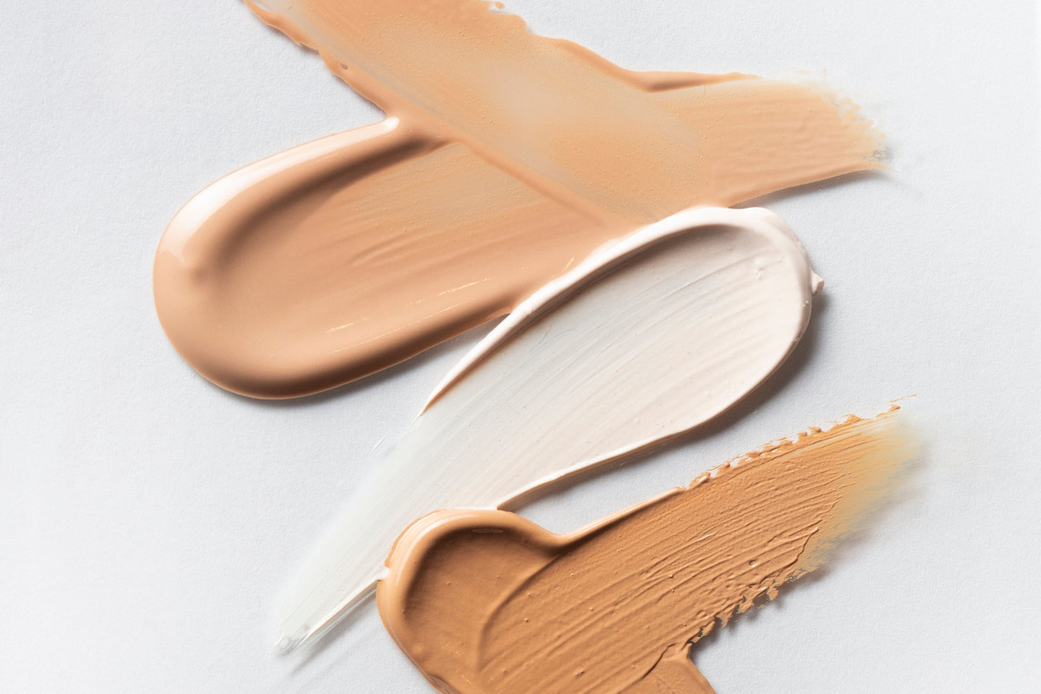 Three swatches of beige makeup cream on a light gray background